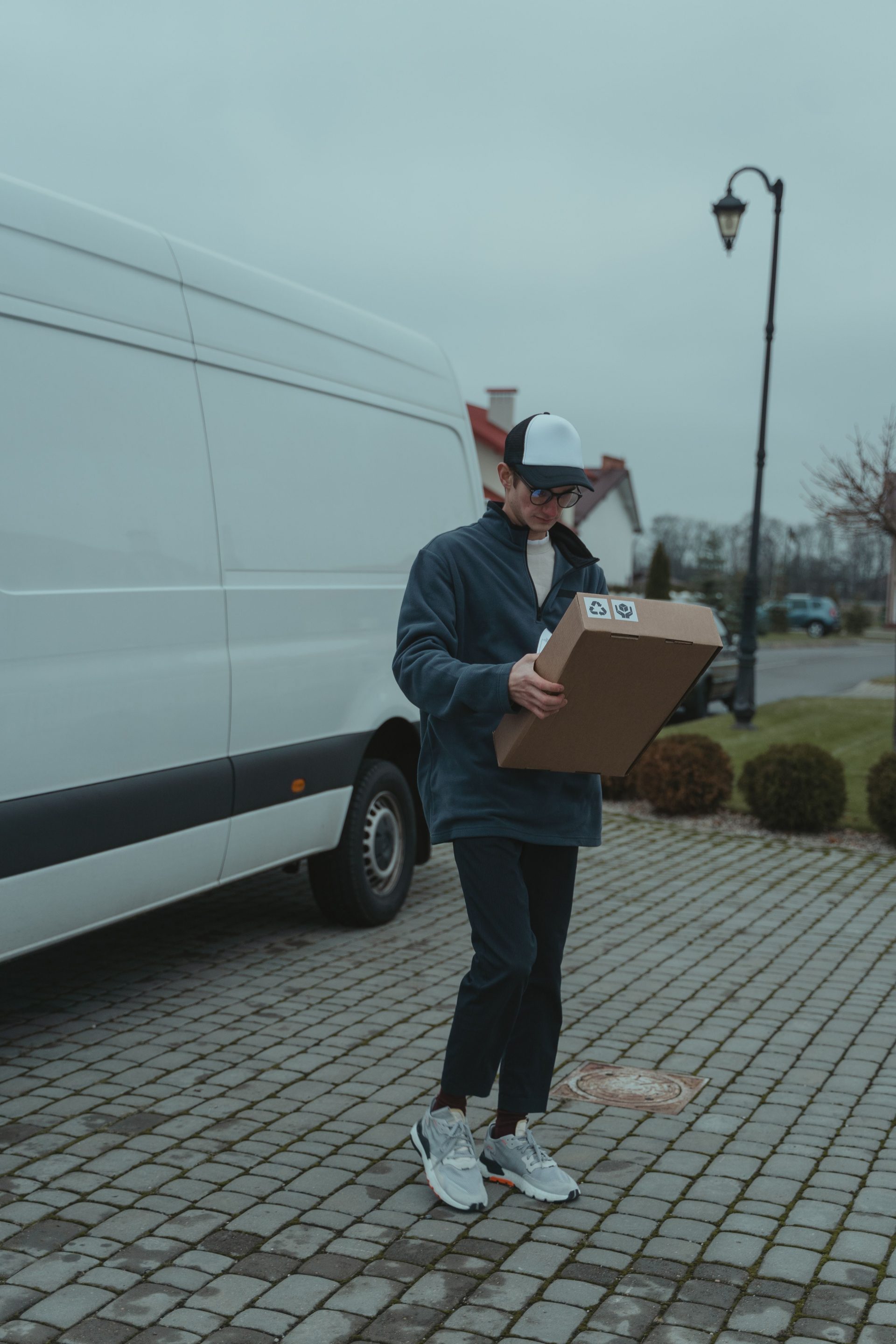 Männlicher Kurier liefert im Freien ein Paket aus; er trägt eine Mütze und eine Jacke und steht neben einem Lieferwagen.