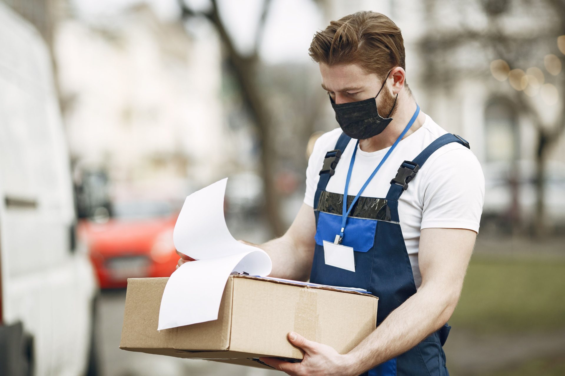 Ein Zusteller mit Gesichtsmaske überprüft im Freien ein Paket. Ideal für Themen aus den Bereichen Logistik und Pandemie.