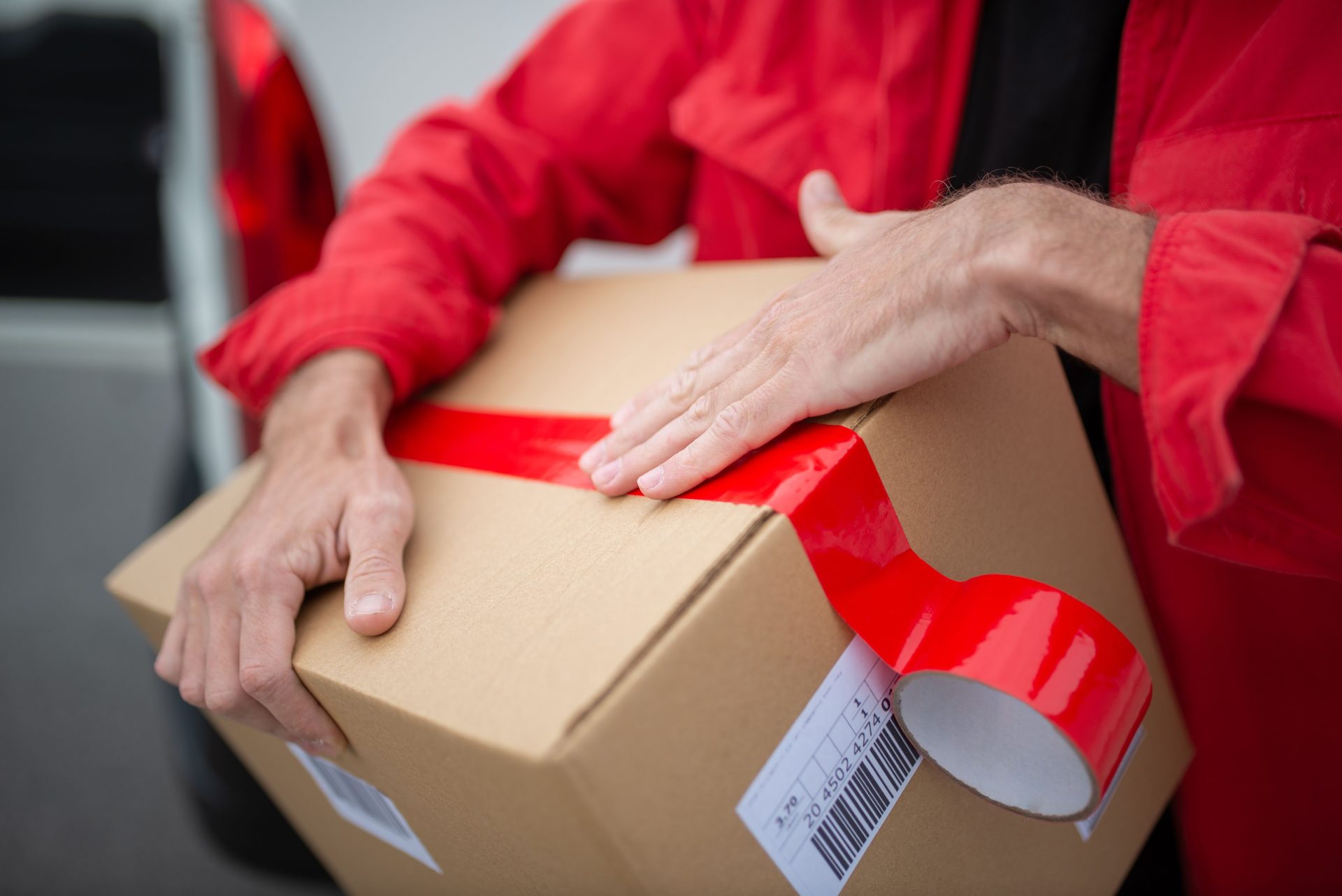 Ein Kurier in roter Uniform verschließt ein Papppaket mit rotem Klebeband für die Zustellung.